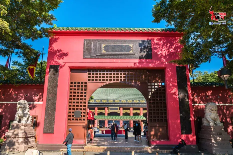 Red temple gate with brown lattice doors leading to a courtyard, visitors entering the complex.