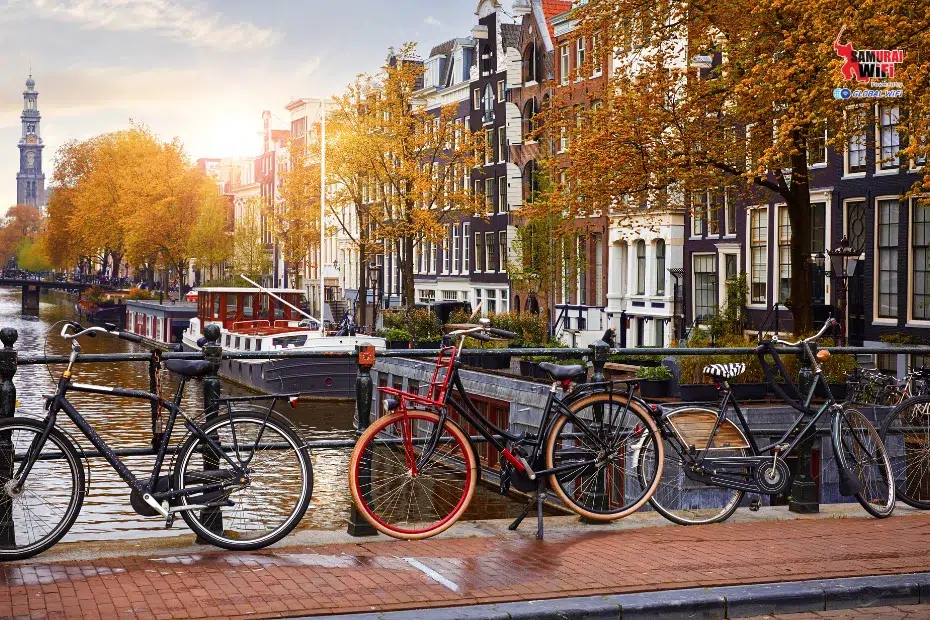 Bikes locked to a railing along a canal in Amsterdam with autumn trees and historic canal houses.