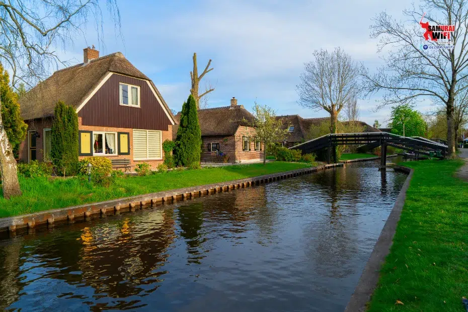Canal lined with thatched-roof cottages, a wooden footbridge, and green grassy banks.