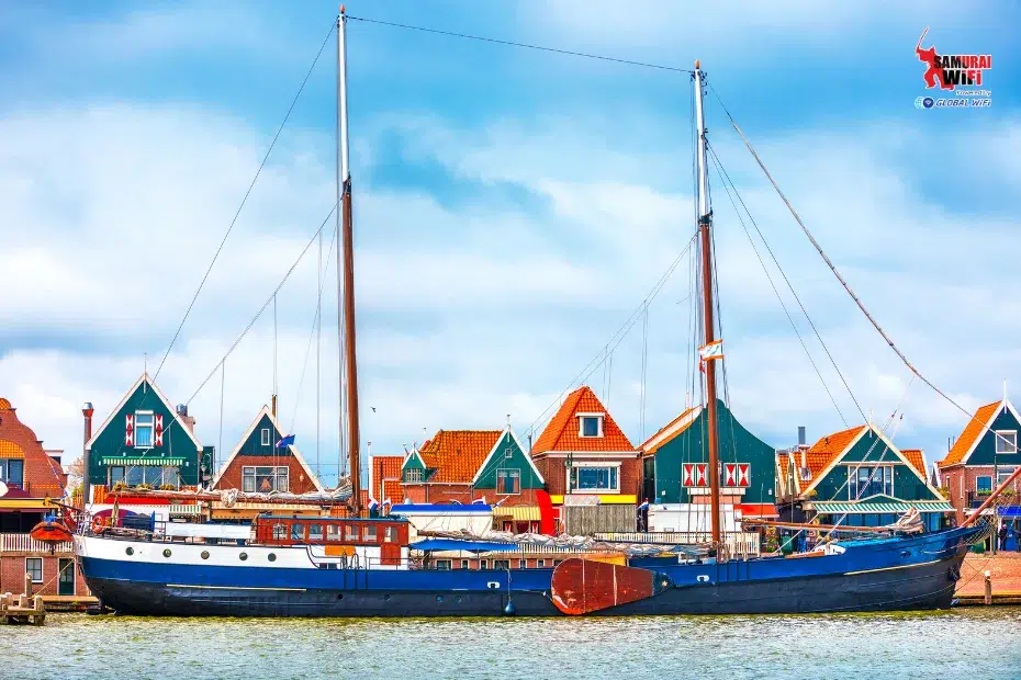 A large blue sailboat docked at a quay with colorful village houses in the background.