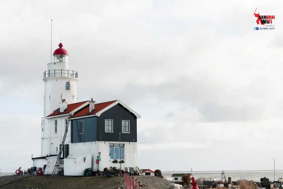 White lighthouse with a red-domed top and attached blue keeper’s house on a rocky coastline under a cloudy sky.