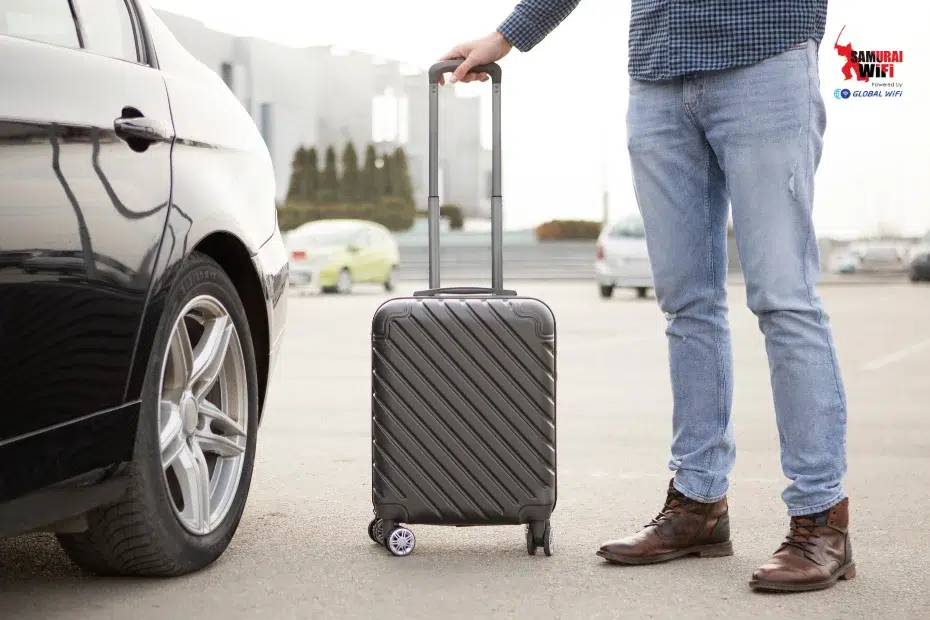 Person in jeans and brown boots stands beside a black rolling suitcase next to a car in a parking lot.
