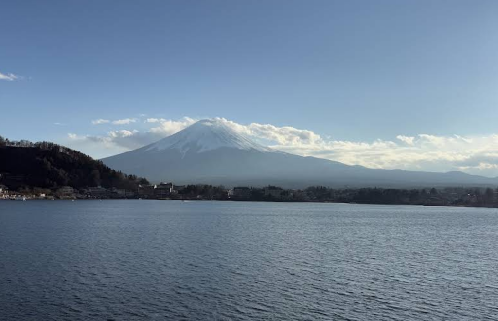 Snow-capped Mount Fuji rises over a calm blue lake with a town along the shoreline beneath a clear sky.