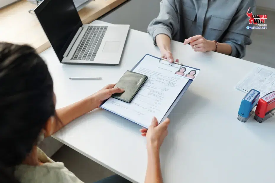 Interviewer reviews a candidate's documents, including a resume on a clipboard and a passport, at a desk with a laptop and stamps nearby. The scene suggests a job or visa interview.